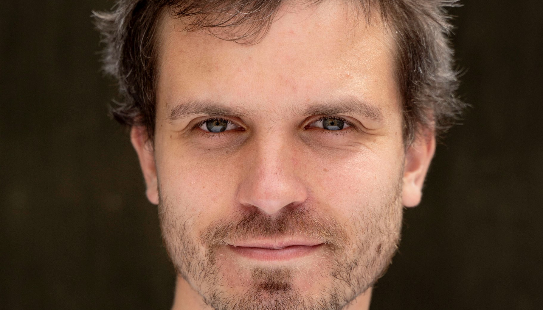 Man med mörkt kort hår, brunt skäggstubb och mustasch. Porträtt. Han tittar rakt in i kamernan