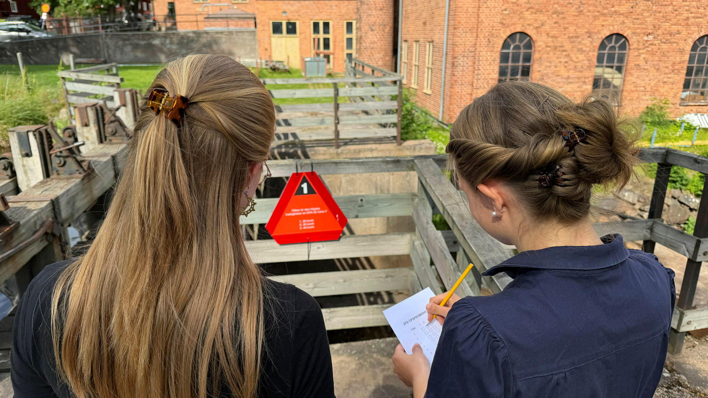 Två personer syns fylla i en något på ett papper, de står vända mot en äldre fabriksbyggnad i tegel