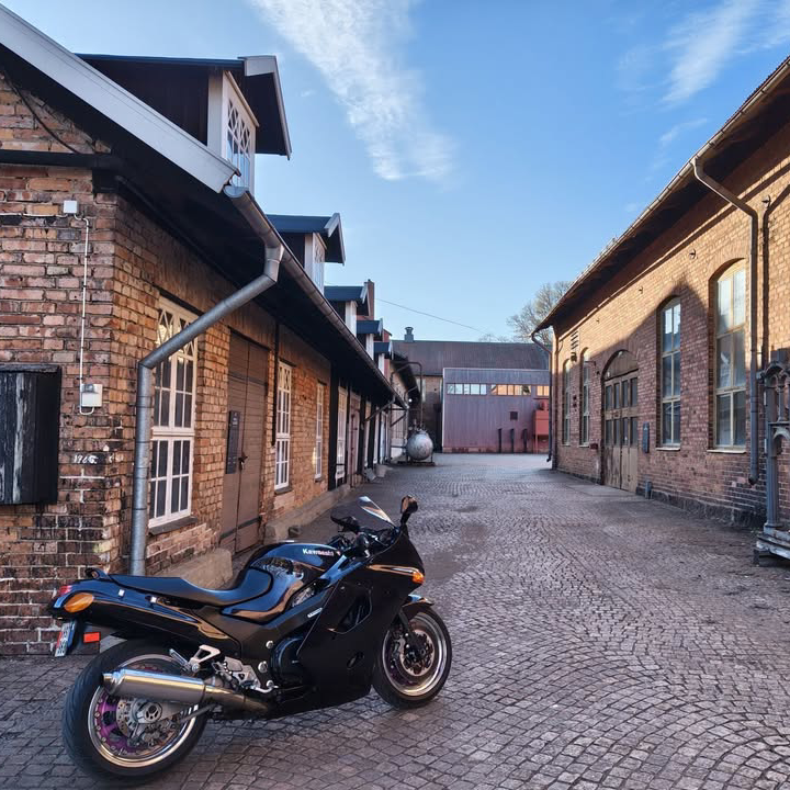 På bilden ser vi en miljö som består av flera tegelbyggnader. Till vänster i bild står en blå motorcykel. 