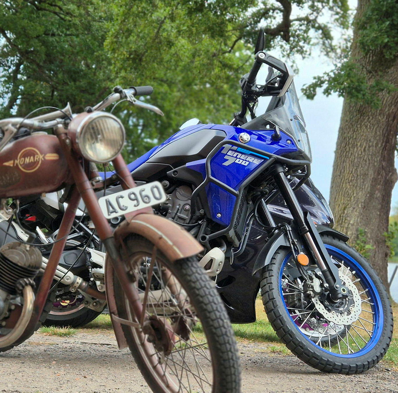 En äldre brun motorcykel står framtill i bild. Bakom den bruna motorcykeln står en något modernare blå motorcykel. I bakgrunden syns träd och en sjö.