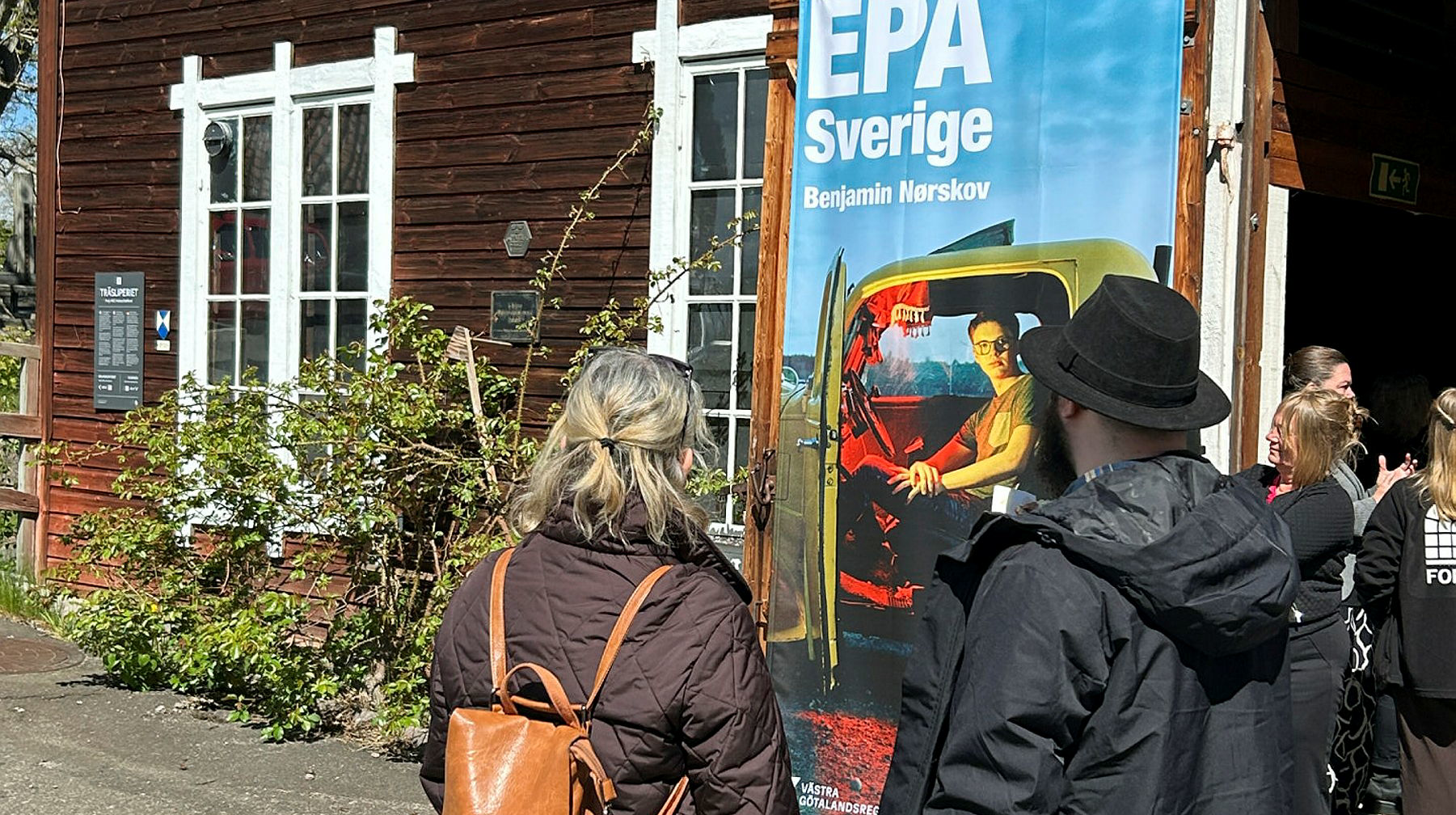 Röd träfasad, fönser med vita fönsterbågar. I närbild, två personer bakifrån.