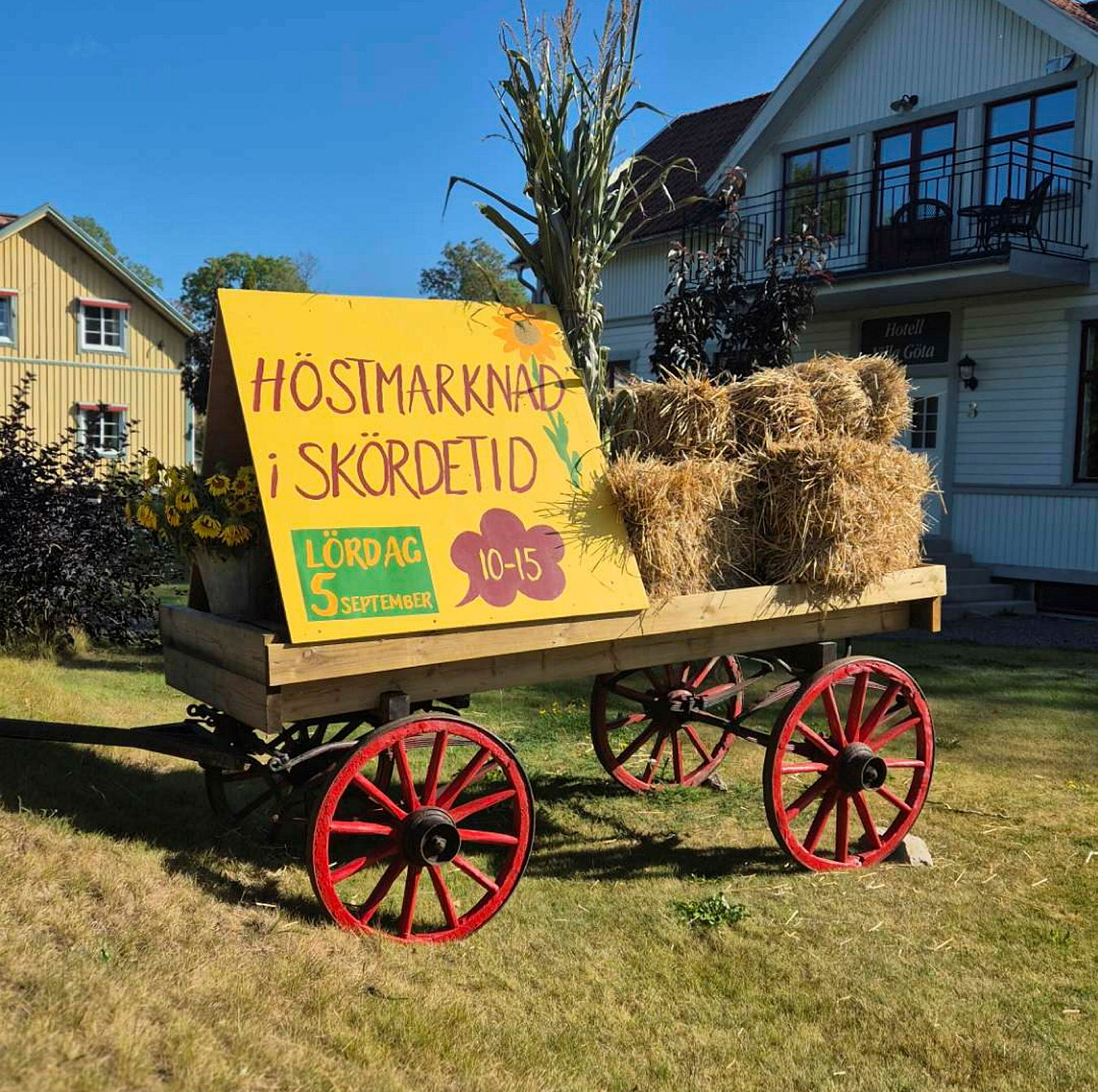 En gammal vagn med röda hjul står på en gräsmatta. På vagnen ligger halmbalar och det står en gul skylt med texten Höstmarknad i skördetid. Lördag 5 september 10-15