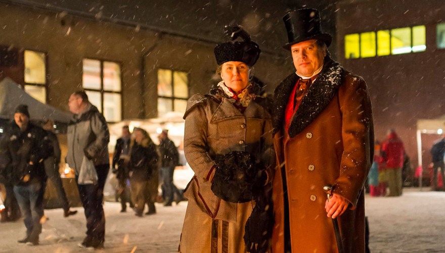 En kvinna och man i rock och hatt från 1900-talets början. Snöväder. I bakgrunden går besökare till julmarknaden och det lyser gult i fönstren i byggnaderna bakom marknadsstånden.