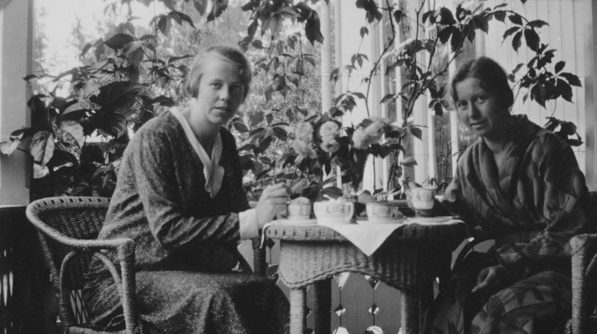 Svartvitt fotografi av två kvinnor som fikar på en altan. De har klänningar på sig och sitter i rottingstolar vid ett tillhörande bord. Bordet är dukat med en vit duk och vita kaffekoppar.