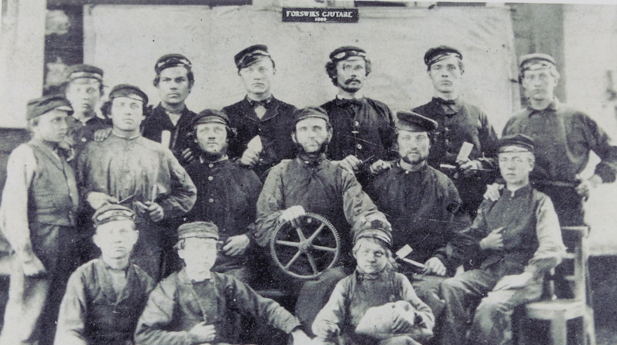 Svartvit bild från 1865 på män och pojkar i arbetskläder som står och sitter i tre rader. Ovanför sitter en svart skylt med texten "Forsviks gjutare 1865".