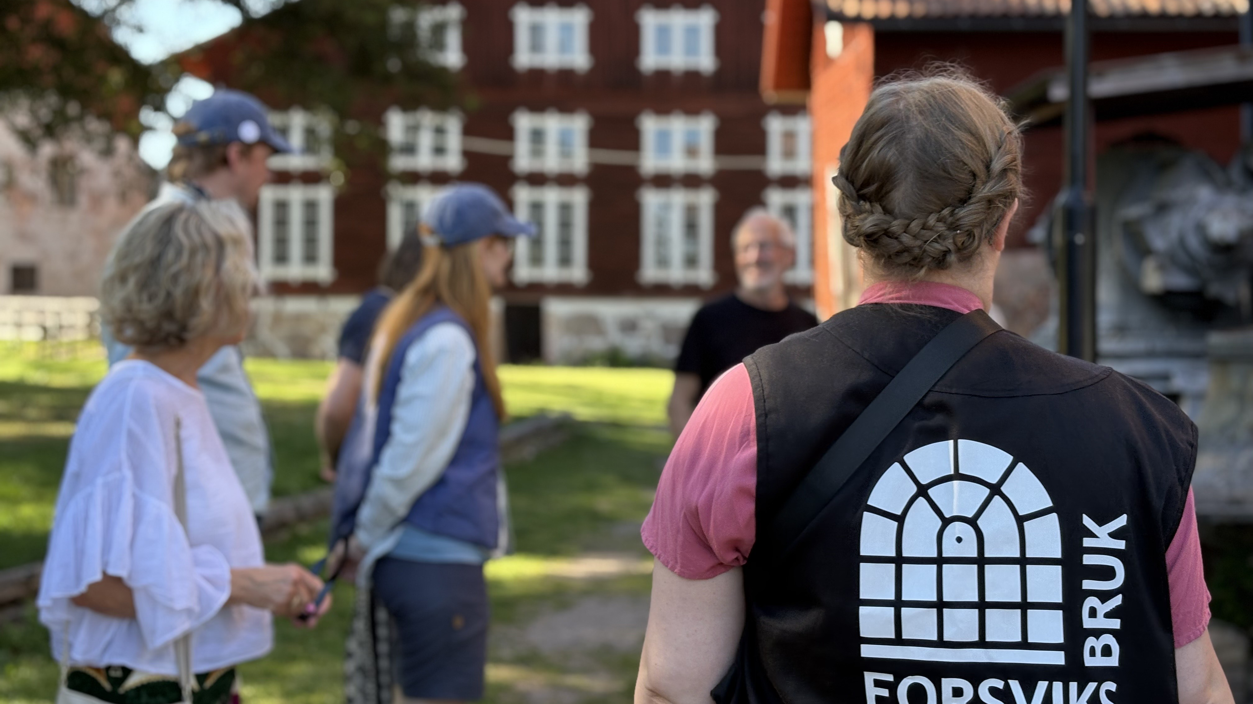 En person med guideväst med Forsviks bruks logga står vänd mot kameran, i bakgrunden en äldre stor träbyggnad samt människor