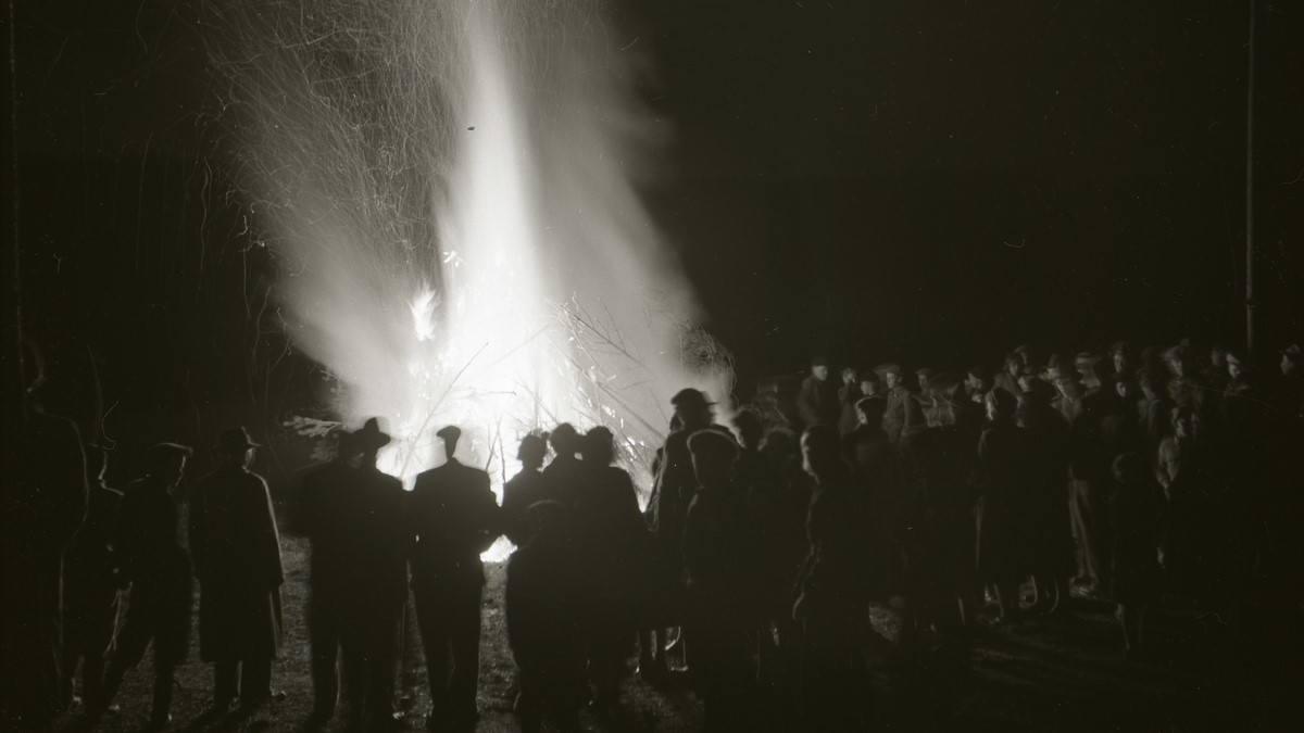 Människor står runt en stor brasa. Kortet är svartvitt, taget 1952.