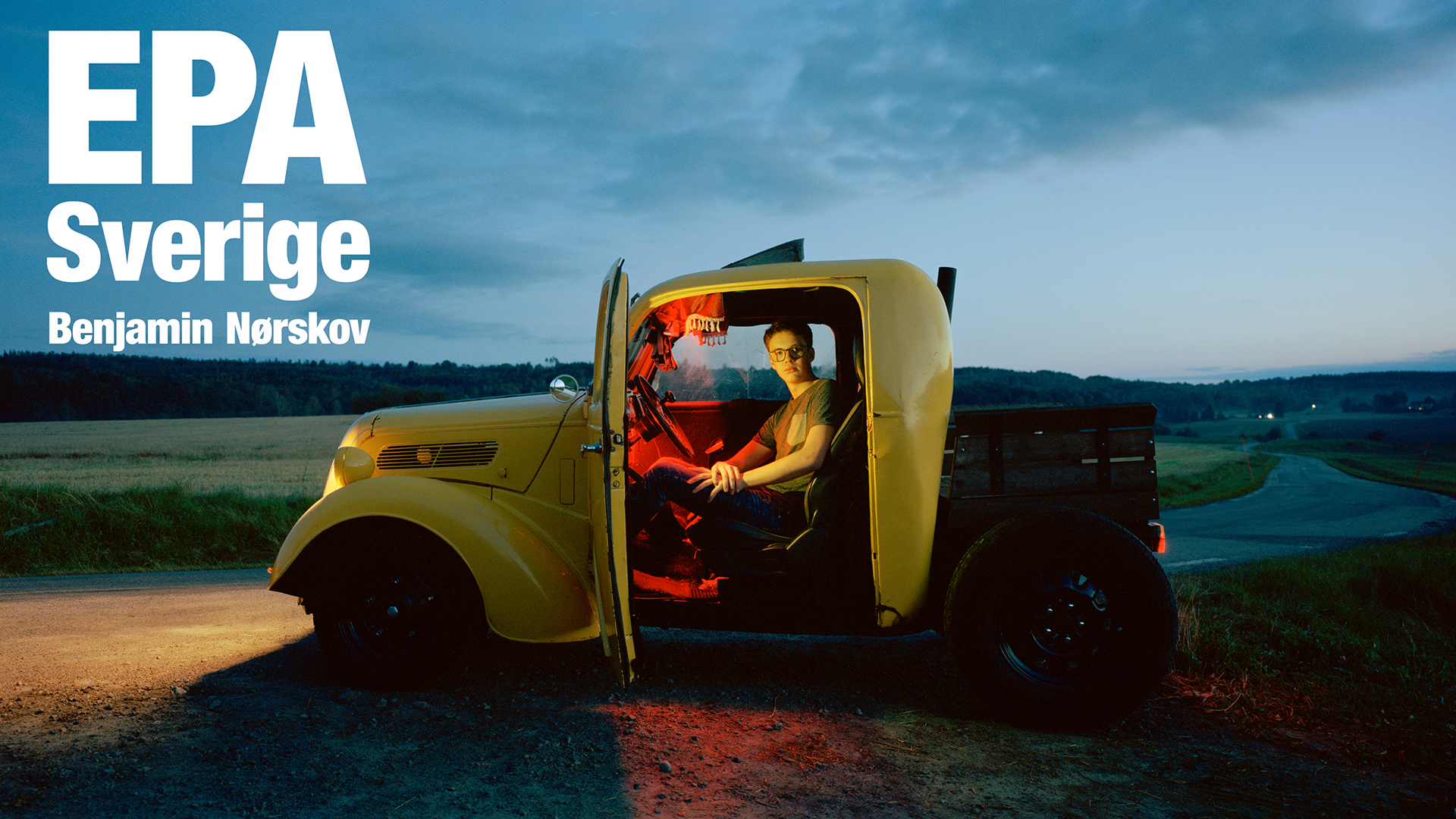 Gul EPA traktor på landsväg. Det är skymning strålkastarna framtill lyser på vägen. Till vänster i bild står det EPA Sverige Benjamin Nörskov i röda bokstäver. I bilen sitter en kille, bildörren till förarsätet är öppen.