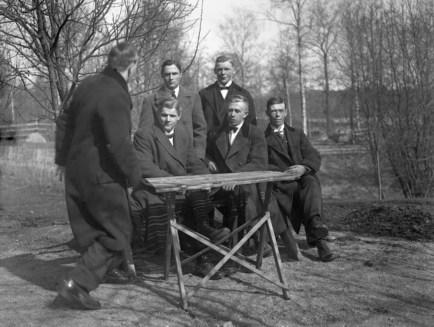 Svartvit bild. Fotografen Sven syns till vänster i bild med ryggen vänd mot kameran. Tre sittande män och två stående män tittar rakt in i kameran. Bild tagen utomhus. Framför de sittande männen står ett trädgårdsbord.